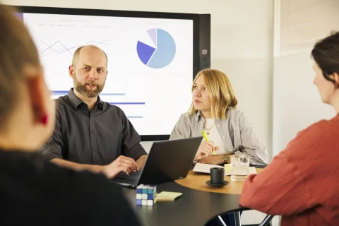 4 Personen reden am Tisch, ein Screen mit Diagrammen im Hintergrund