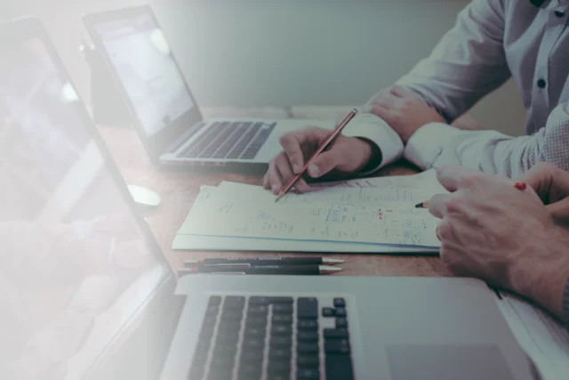 Hände am Schreibtisch mit Dokumenten und Laptops