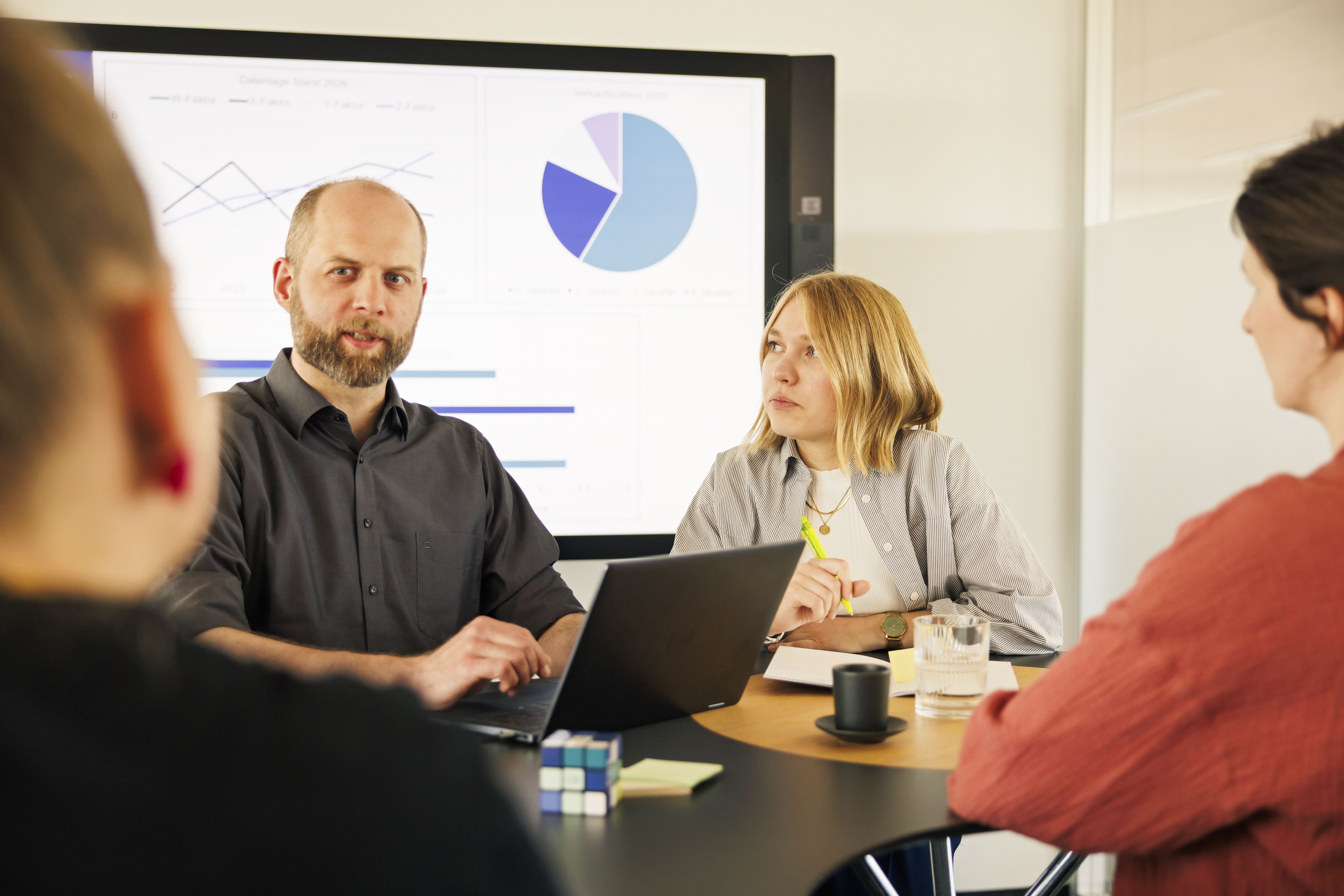 4 Personen reden am Tisch, ein Screen mit Diagrammen im Hintergrund